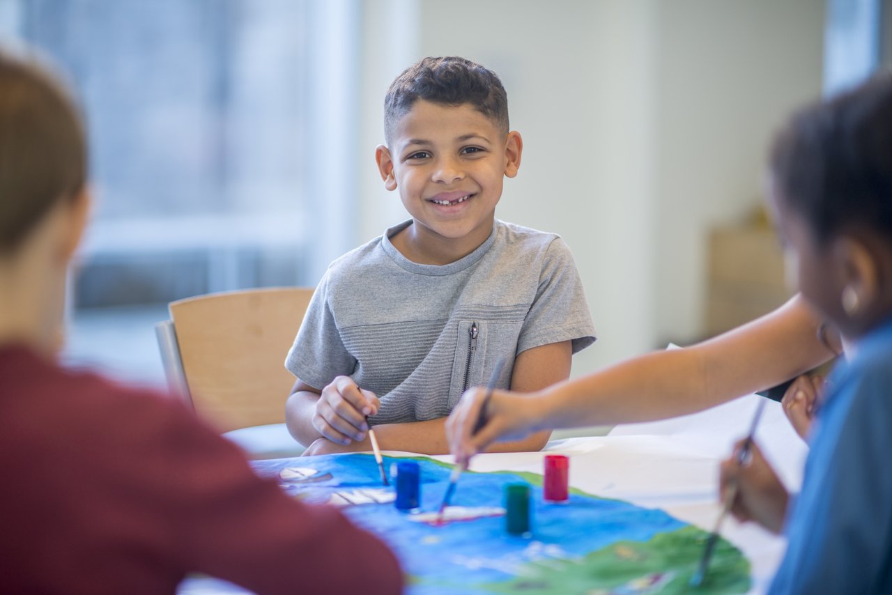 Aluno em aula de arte faz pinturas em uma mesa junto com outras crianças