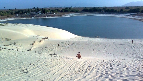 Duna de areia branca que termina em um lago azul com uma pessoa ao centro da foto escorregando rumo ao lago