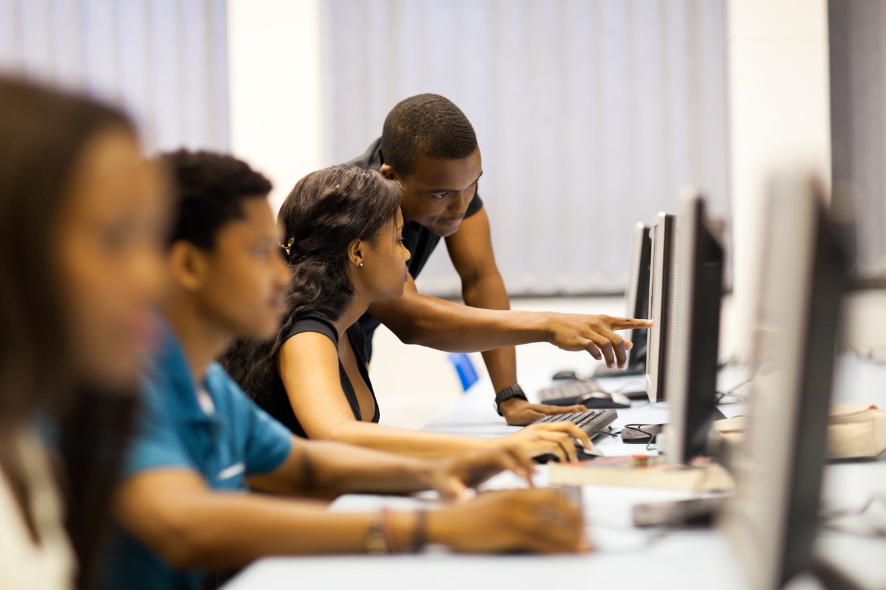 Grupo de alunos em frente a monitores de computadores e professor ao fundo, ajudando uma aluna