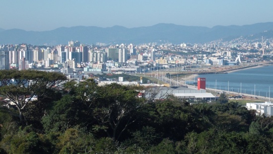 ao fundo, uma cordilheira de montanhas quase se mistura com o céu. Nota-se uma grande quantidade de prédios, algumas empresas industriais e no primeiro plano algumas árvores. No canto direito, há o caminho da orla e do mar por toda a cidade.