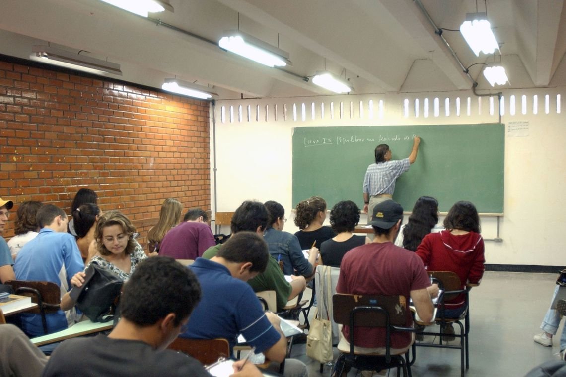Alunos de Ensino Médio sentados de costas para a câmera em uma sala de aula onde o professor escreve em uma lousa verde