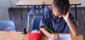 um menino de origem asiática com a cabeça abaixada e mão em um dos olhos. Ele está sentado em uma carteira escolar, com um caderno e segurando um lápis e usa máscara.