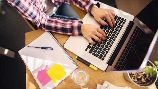 mesa de trabalho vista de cima com notebook aberto e caderno aberto com post-its colados em uma folha em branco. É possível ver os braços de uma mulher usando camisa xadrez que parece estar mexendo no computador.