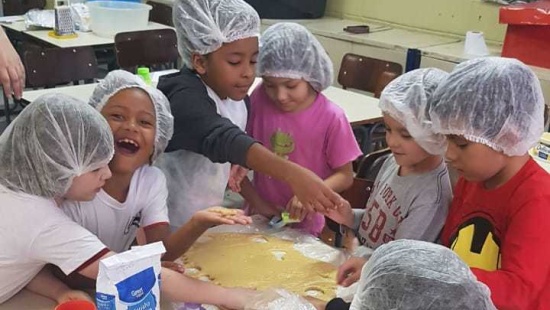 Crianças de touca higiênica na cabeça mexem em uma forma com massa de biscoito espalhada. Uma das crianças olha para a frente da foto e sorri