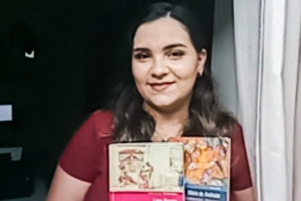 Fotografia produzida por vídeo chamada com a professora Marcia. A personagem segura alguns livros em sua janela.
