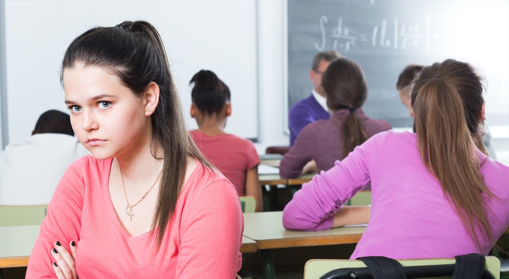 Jovem com expressão triste veste blusa rosa virada de frente para a imagem mas de costas para grupo reunido em sala de aula ao fundo
