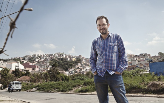 Retrato do professor Fábio Augusto Machado vestindo camisa xadrez e com as mãos nos bolsos da calça jeans. Ao fundo, vê-se a comunidade que o professor leciona.