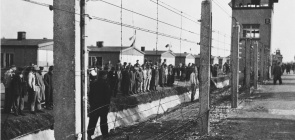 Na foto, em branco e preto, vemos uma cena de um grupo de pessoas atrás de uma cerca. Trata-se de um campo de concentração em Dachau