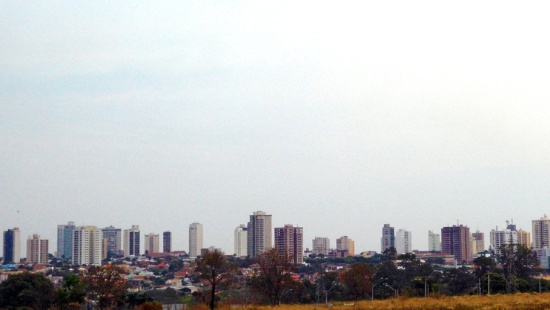 nuvens brancas encobrem todo o céu. Na linha horizontal na parte inferior da imagem, um gramado verde com árvores ao fundo e prédios dispostos atrás dessas árvores