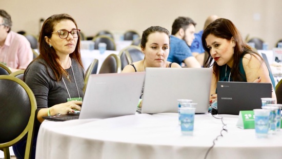 três professoras, cada uma com seu laptop, sentadas ao lado uma da outra diante de uma mesa branca (onde os computadores estão abertos)