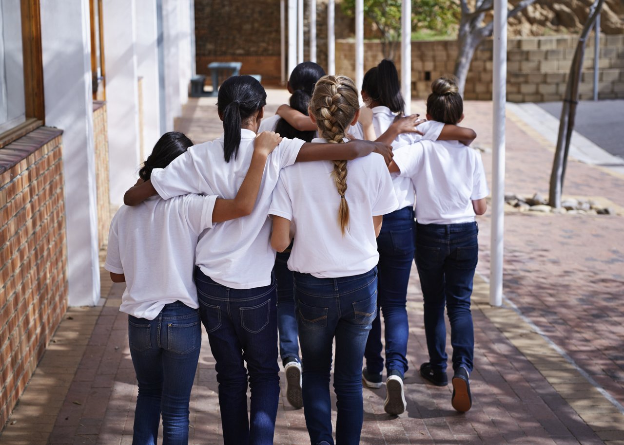Adolescentes vestindo calças jeans e camisetas brancas caminham abraçadas perto de um muro de tijolos aparentes