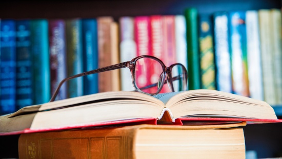 Óculos em cima de livro aberto com uma parede cheia de livros atrás