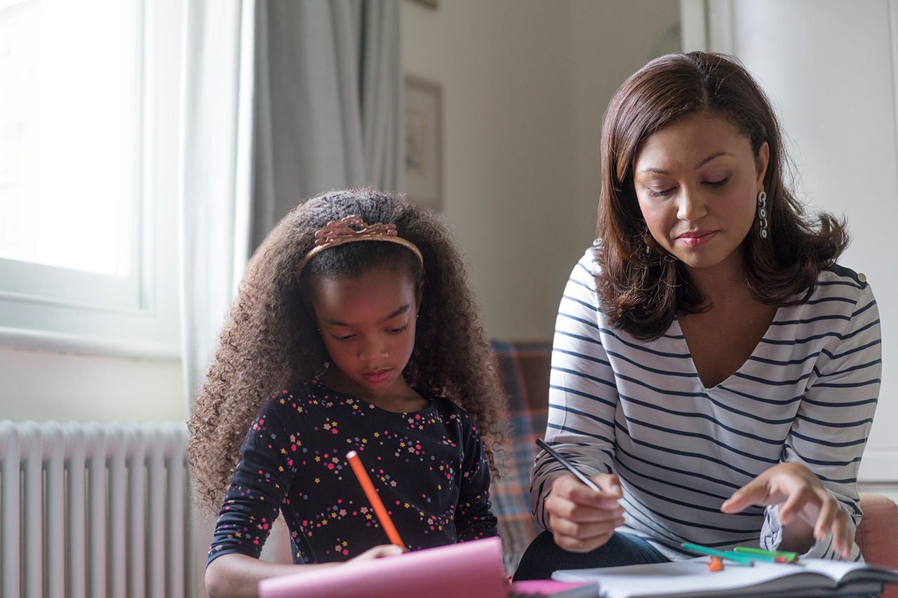 Mãe e filha estudando juntas com livros e cadernos sobre mesa. A mãe auxilia a filha no processo de alfabetização.