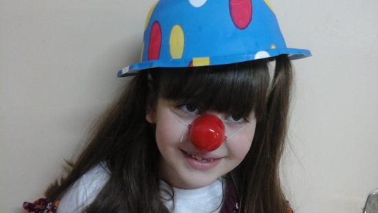 menina branca de longos cabelos castanhos e lisos e franja sorri vestindo um chapéu colorido (azul com bolas rosas) e nariz de palhaço