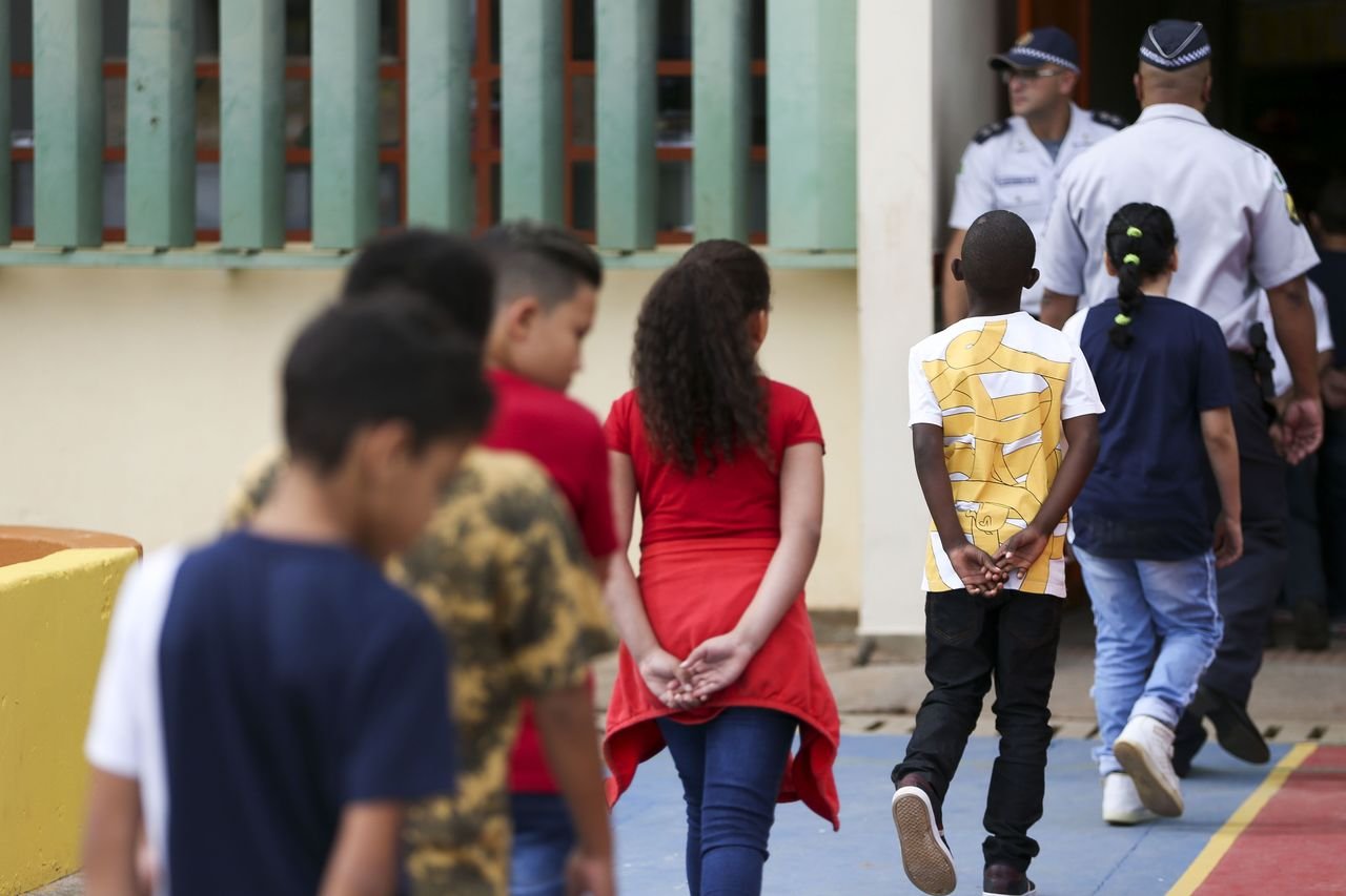 Crianças de costas para a câmera caminham para a entrada da escola no CED 01 da Estrutural, uma das escolas públicas do DF onde foi implementado o modelo cívico-militar. Policiais militares observam a entrada na porta