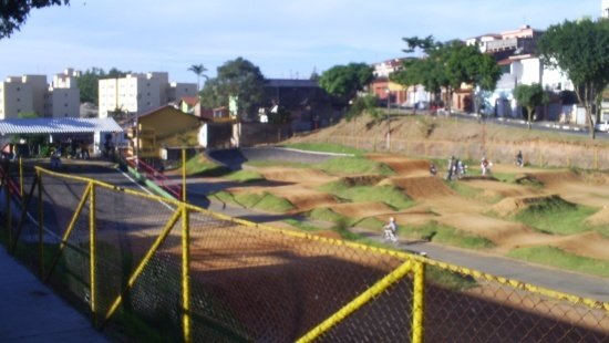 O primeiro plano mostra uma grade amarela. Atrás dela, revela-se uma área verde coberta por grama com ondulações no solo.