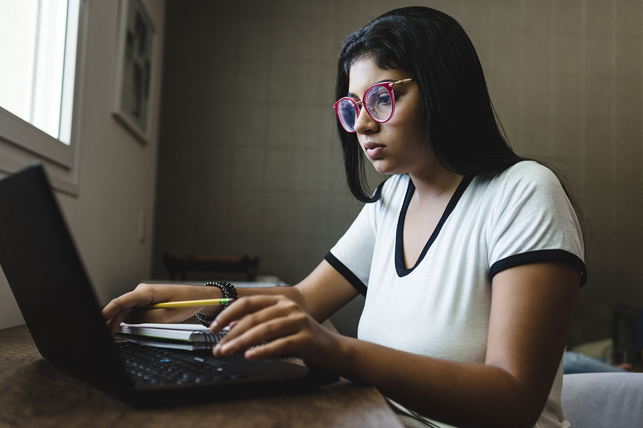 Fotografia de aluna do ensino médio em frente ao computador fazendo anotações em seu caderno.