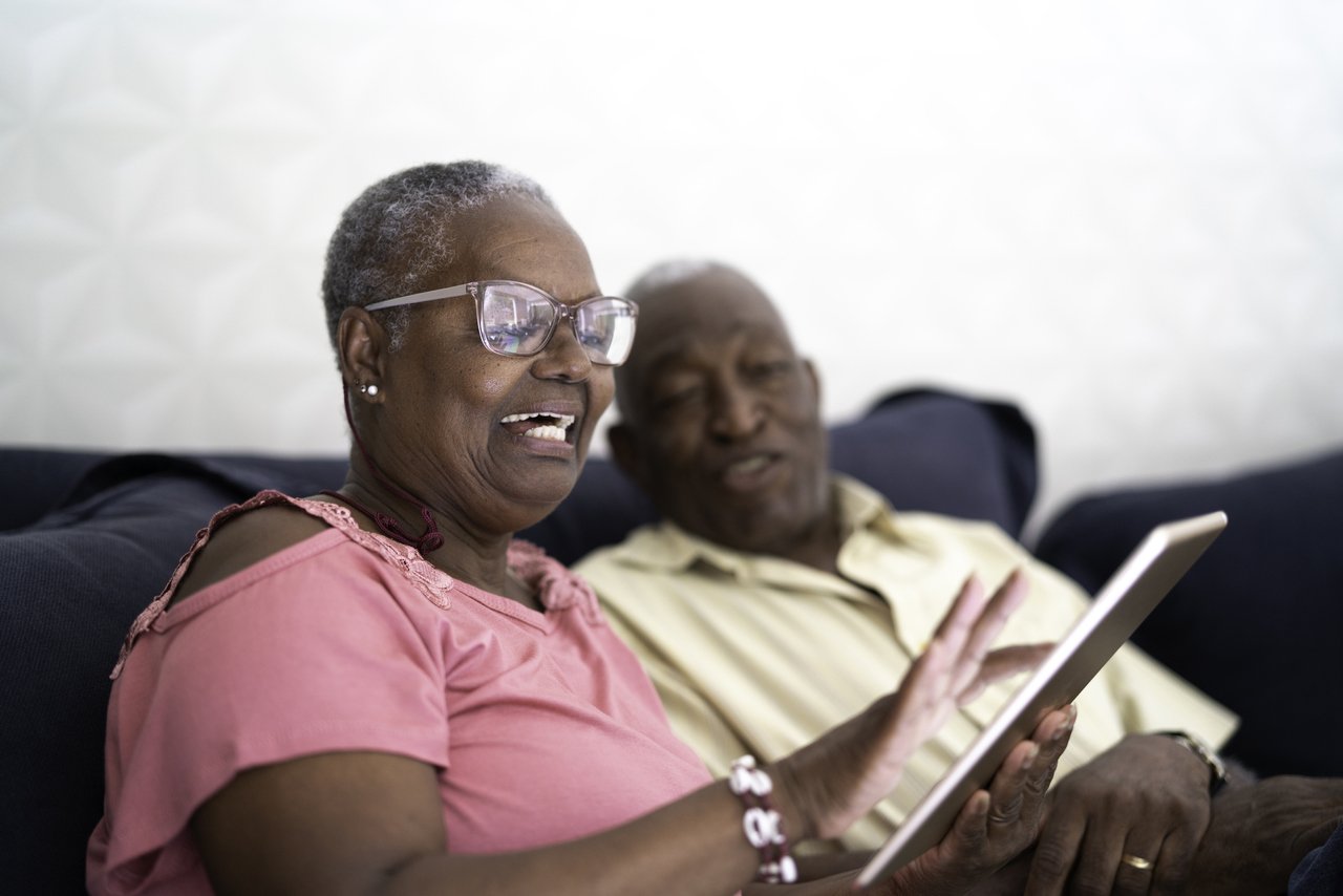 senhora negra com tablet na mão e ao fundo da foto um senhor também observando o tablet