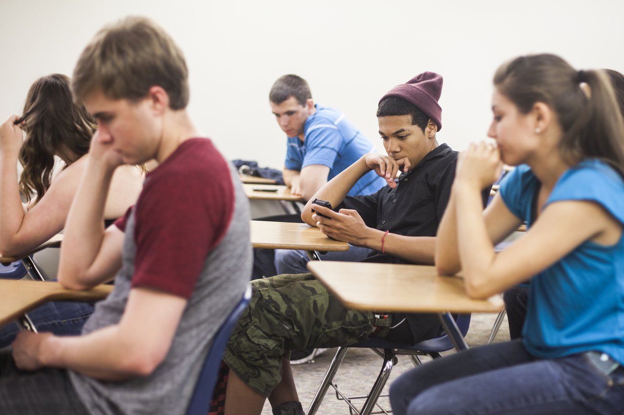 Em uma sala de aula, em meio aos alunos, um estudante não presta atenção na aula e olha para o celular