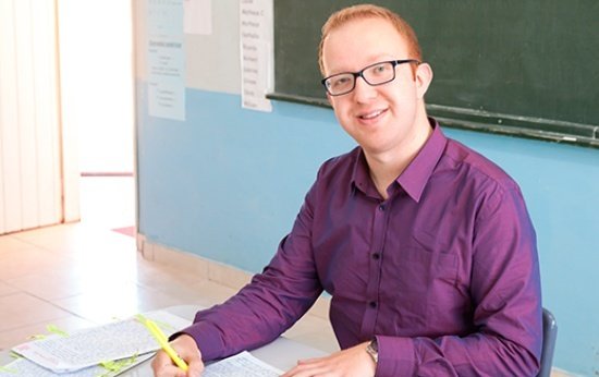 Relato do educador nota 10 Carlos Eduardo Canani sentado em sua mesa na sala de aula. Sobre a mesa estão diversos papéis e cadernos.