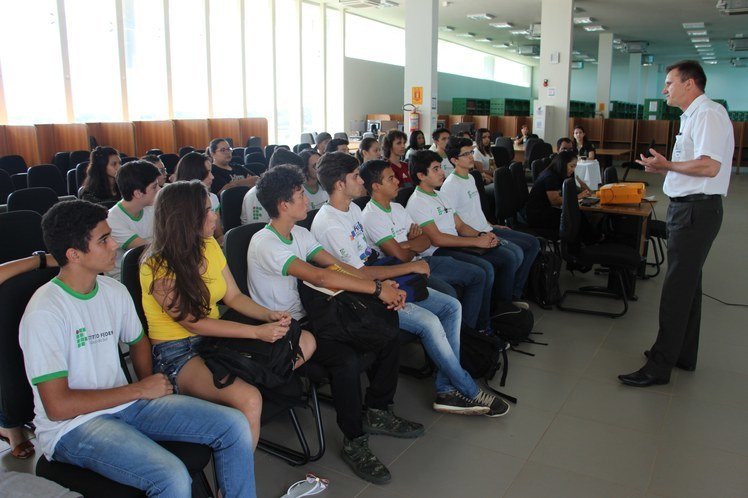 Fila de jovens sentados em cadeiras ouvindo professor falar à frente, de pé