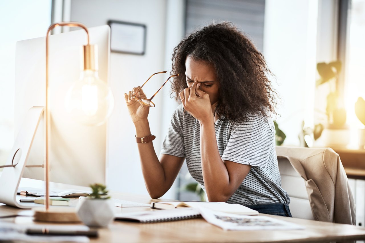 Mulher sentada em uma mesa em frente a um monitor de computador tira os óculos e segura entre os olhos com uma das mãos em uma expressão de cansaço