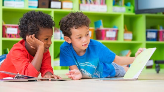 Dois meninos, um de camiseta vermelha e outro de camiseta azul, estão deitados no chão da sala de aula. O de camiseta azul folheia um livro enquanto o de camiseta vermelha apoia a cabeça na mão.