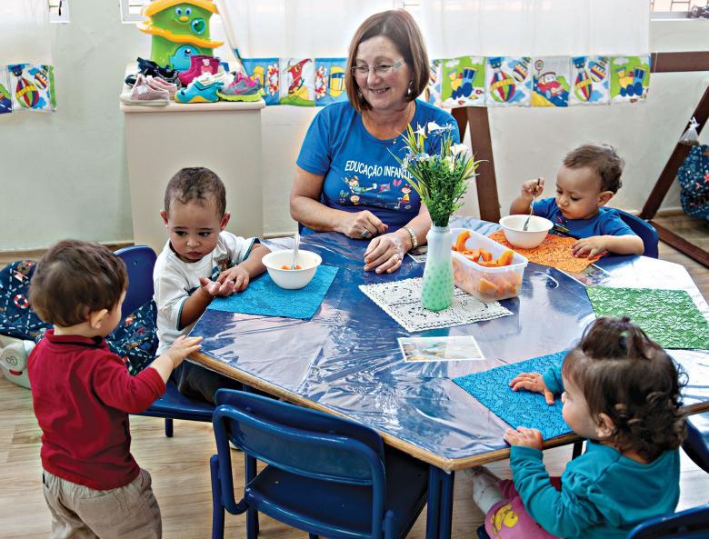 Professora e crianças na mesa do almoço do NEI Otilia Cruz