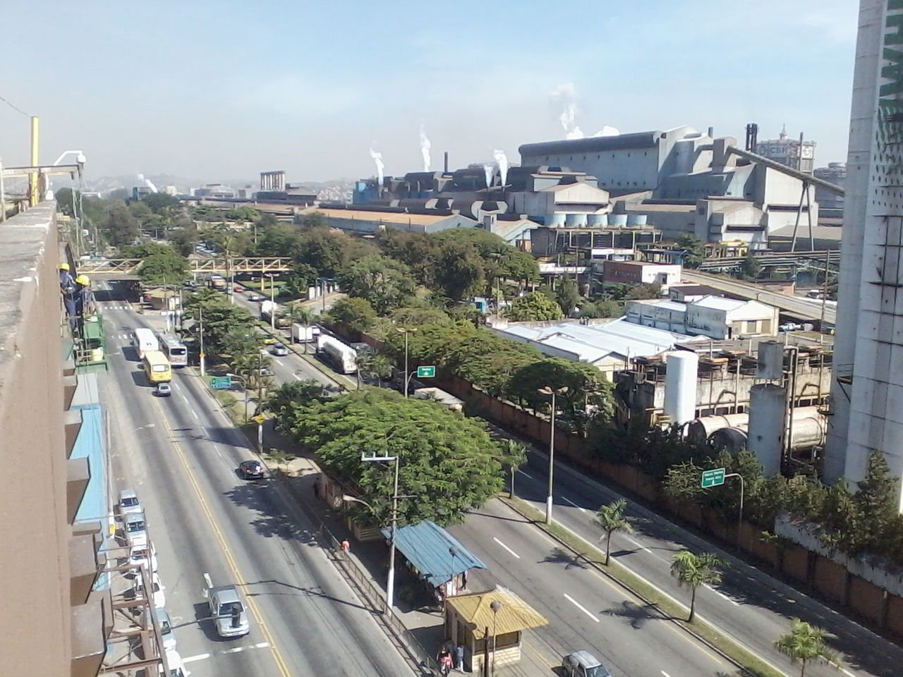 Avenida movimentada vista lateralmente e de cima com fábricas soltando fumaça de suas chaminés e carros passando na pista